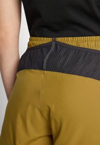 Stretchy athletic pants featuring a dark gray, textured mesh waistband and a mustard yellow body with subtle vertical ribbing.