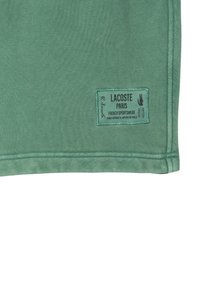 Tissu en coton vert avec un ourlet cousu. Présente une étiquette bleue avec un texte noir indiquant "Lacoste Paris French Sportswear."