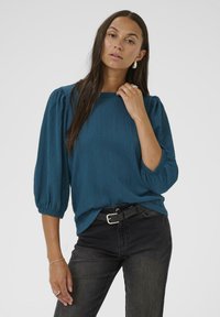 Haut texturé teal avec des manches bouffantes et un col rond, associé à un jean noir et une ceinture noire. Cheveux longs et accessoires minimalistes.