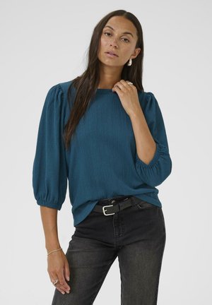 Teal textured top with puffed sleeves and a crew neckline, paired with black jeans and a black belt. Long hair and minimal accessories.