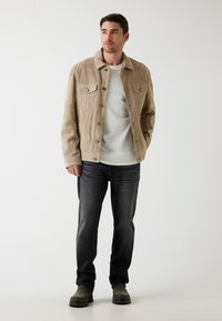Beige Wildlederjacke mit kragenlosem Hals, vorderen Druckknöpfen und zwei Brusttaschen, kombiniert mit einem weißen Hemd und dunklen Jeans.
