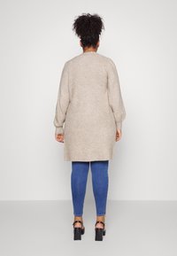 Cardigan beige lungo con scollo rotondo, polsini a coste e silhouette leggermente affusolata, abbinato a jeans skinny blu e sandali neri con tacco a blocco.