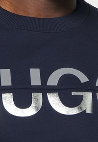 Primo piano di una felpa girocollo blu navy con grandi lettere metalliche argento che formano parzialmente una parola sul petto.