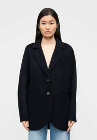 Mustast villa segu segisti tippkraega, ühe nupuga kinnitusega ja kahe eest taskuga; sile tekstuur, struktureeritud lõige, puusapikkune disain.