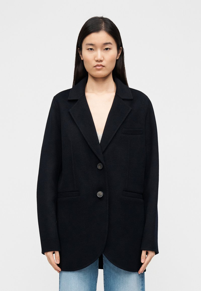 Mustast villa segu segisti tippkraega, ühe nupuga kinnitusega ja kahe eest taskuga; sile tekstuur, struktureeritud lõige, puusapikkune disain.