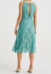 Teal lace dress with scalloped hem and open back, featuring a tie closure. Designed with a fitted bodice and flared skirt.