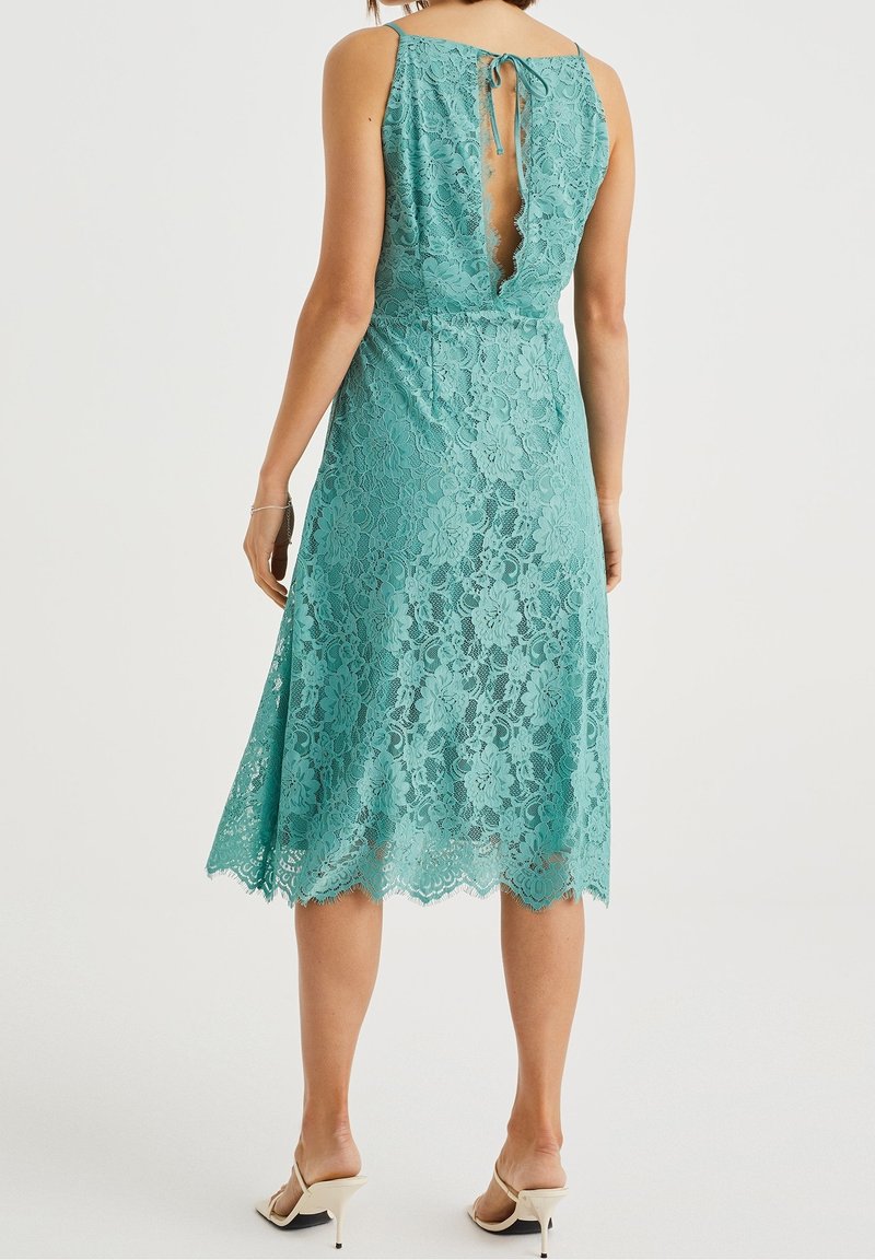 Teal lace dress with scalloped hem and open back, featuring a tie closure. Designed with a fitted bodice and flared skirt.