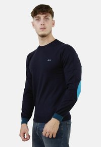 Maglione blu navy a maniche lunghe realizzato in tessuto liscio, con un'accents turchese sulla manica sinistra e un piccolo logo sul petto.
