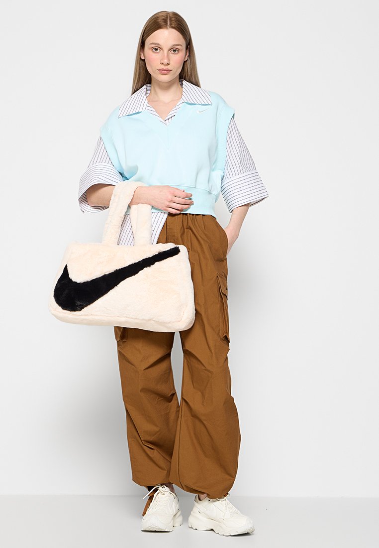 Borsa in pelliccia color crema con logo Nike nero, forma rettangolare e due manici corti. Il modello indossa un top corto blu chiaro e pantaloni cargo marroni.