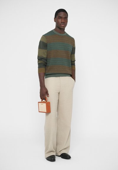 Sweter dziergany z teksturowanymi zielonymi i brązowymi paskami, beżowe spodnie oraz mała drewniana torebka z brązowym uchwytem. Całość uzupełniają czarne buty.