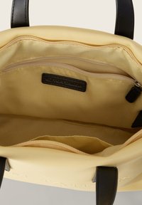 Geel canvas tas met zwarte handvatten, voorzien van een ritsvak aan de binnenkant en een hoofdcompartiment gevoerd met zachte stof. Merketiket zichtbaar aan de binnenkant.