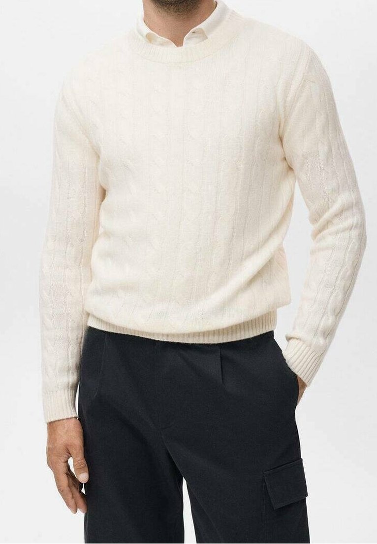Homme portant un pull en maille torsadée de couleur blanc cassé et un pantalon noir à coupe décontractée avec une poche latérale, debout avec une main dans la poche.