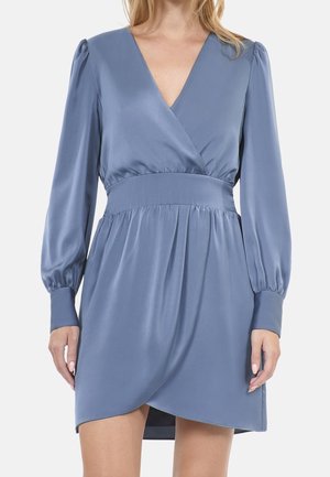 Kobieta ubrana w długą, jasnoniebieską satynową sukienkę kopertową z głębokim dekoltem w kształcie litery V i dopasowaną talią.