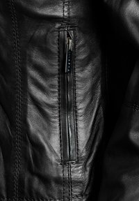 Dettaglio di una giacca in pelle nera con una tasca con zip, texture liscia, cuciture visibili ed elementi di design eleganti.