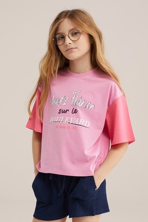 Jong meisje met lang blond haar en een bril, draagt een roze t-shirt met Franse tekst en marineblauwe korte broek, handen in de zakken, neutrale achtergrond.