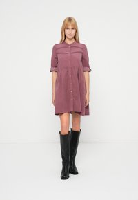 Lawendowa sukienka zapinana na guziki, z marszczonym pasem, krótkimi podwiniętymi rękawami i delikatnym teksturowanym wykończeniem na wysokości klatki piersiowej. Noszona z czarnymi kozakami za kolano.