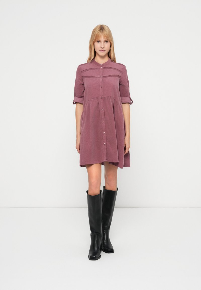 Lawendowa sukienka zapinana na guziki, z marszczonym pasem, krótkimi podwiniętymi rękawami i delikatnym teksturowanym wykończeniem na wysokości klatki piersiowej. Noszona z czarnymi kozakami za kolano.