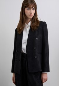 Sort blazer med tynde striber, dobbeltbrystet forside og store knapper, kombineret med en hvid skjorte med krave. Glat stoftekstur, struktureret design.