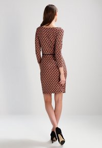 Dopasowana sukienka z geometrycznym czerwono-czarnym wzorem, rękawami do połowy łokcia i wyraźnie zaznaczoną talią. Noszona z czarnymi butami na wysokim obcasie.