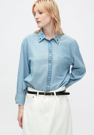 Donna che indossa una camicia di jeans ricamata blu chiaro, infilata in una gonna bianca a vita alta con cintura nera, mentre guarda di lato.