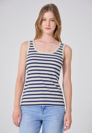Femme aux cheveux clairs portant un haut sans manches crème et bleu marine à rayures et un jean bleu clair, debout sur un fond uni.