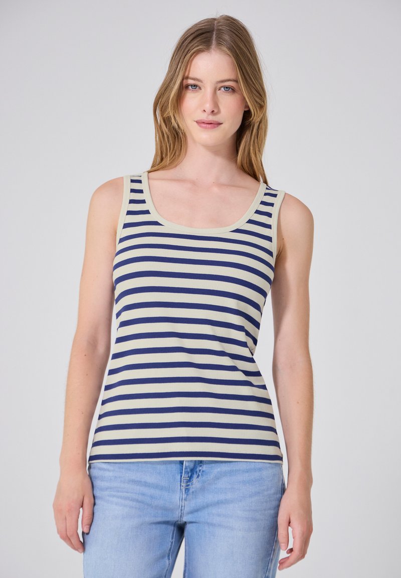 Femme aux cheveux clairs portant un haut sans manches crème et bleu marine à rayures et un jean bleu clair, debout sur un fond uni.
