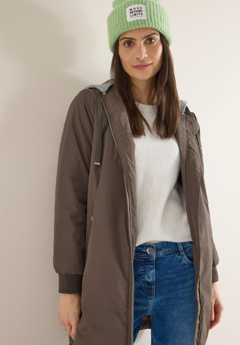 Femme portant un bonnet vert avec le texte « Move Beyond Limits », une veste marron zippée, un pull blanc et un jean bleu, debout contre un mur uni.