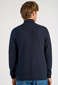 Marinblå stickad tröja med hög krage, texturerat mönster och ribbade manschetter. Tyget är mjukt och figurnära vid midjan.