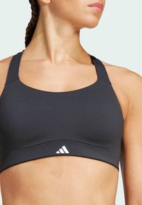 Czarny stanik sportowy z gładkiego materiału, z zaokrąglonym dekoltem, szerokimi ramiączkami i białym logo w pobliżu dolnej krawędzi.
