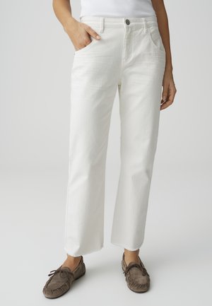 Persoon draagt witte high-waist rechte jeans met onafgewerkte zomen, één hand in de zak, gecombineerd met bruine mocassins.