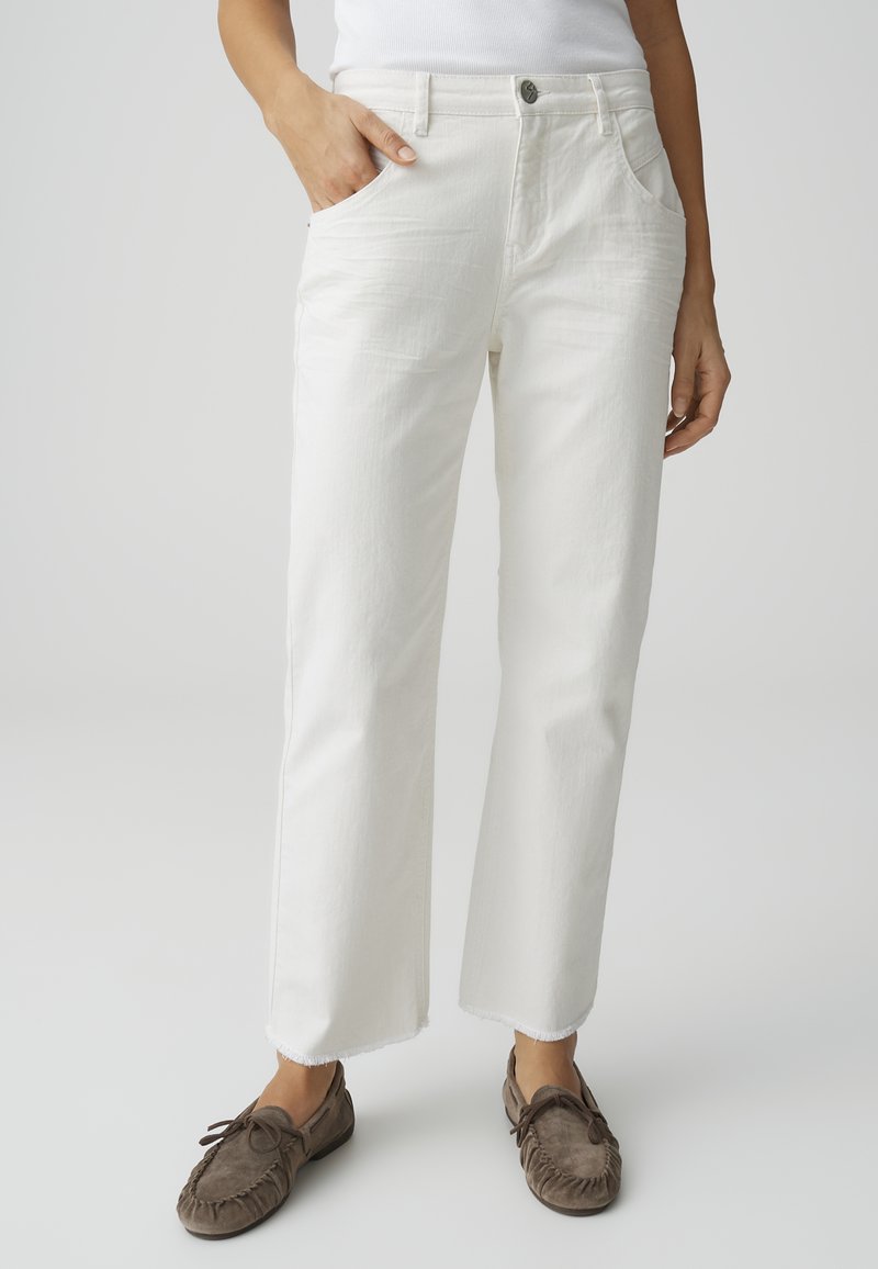 Person wearing high-waist white straight-leg jeans with raw hems, one hand in pocket, paired with brown moccasin shoes.