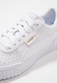 Zapatilla blanca de cuero con superficie texturizada, puntera redonda, perforaciones y cordones blancos. Presenta un logo dorado de Puma en el lateral.