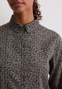 Zwart overhemd met bloemenpatroon, een kraag, knopen aan de voorkant en lange mouwen. De stof heeft een zachte textuur en kleine, delicate witte bloemen.