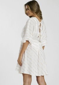 Vestido blanco con rayas doradas, de corte holgado y manga corta. Se ata en la parte posterior del cuello y tiene un dobladillo con escamas. Tejido ligero con apariencia texturizada.