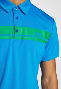 Pánské polokošile v jasně modré barvě s výrazným zeleným horizontálním pruhem. Má klasický límec a tříknoflíkovou légu. Hladká, lehká látka.