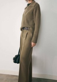 Khaki žebrovaný pletený svetr s límečkem, doplněný o kalhoty s vysokým pasem a širokými nohavicemi. Černá kabelka přidává kontrast. Převládají neutrální tóny.