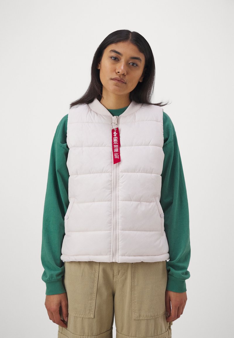 Puffweste ohne Ärmel in hellrosa, mit einem Reißverschluss vorne, seitlichen Taschen und einem roten Etikett. Getragen über einem grünen Hemd und beigen Hosen.