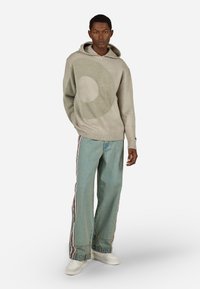 Férfi áll beige kapucnis pulóverben, amelyen kör alakú minta látható, széles lábú kék farmerben csíkos oldalakkal, valamint fehér tornacipőben, fehér háttér előtt.