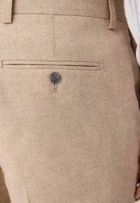 Los pantalones de lino beige cuentan con una tela texturada con un sutil tejido, un bolsillo delantero y un único botón gris en la parte posterior.