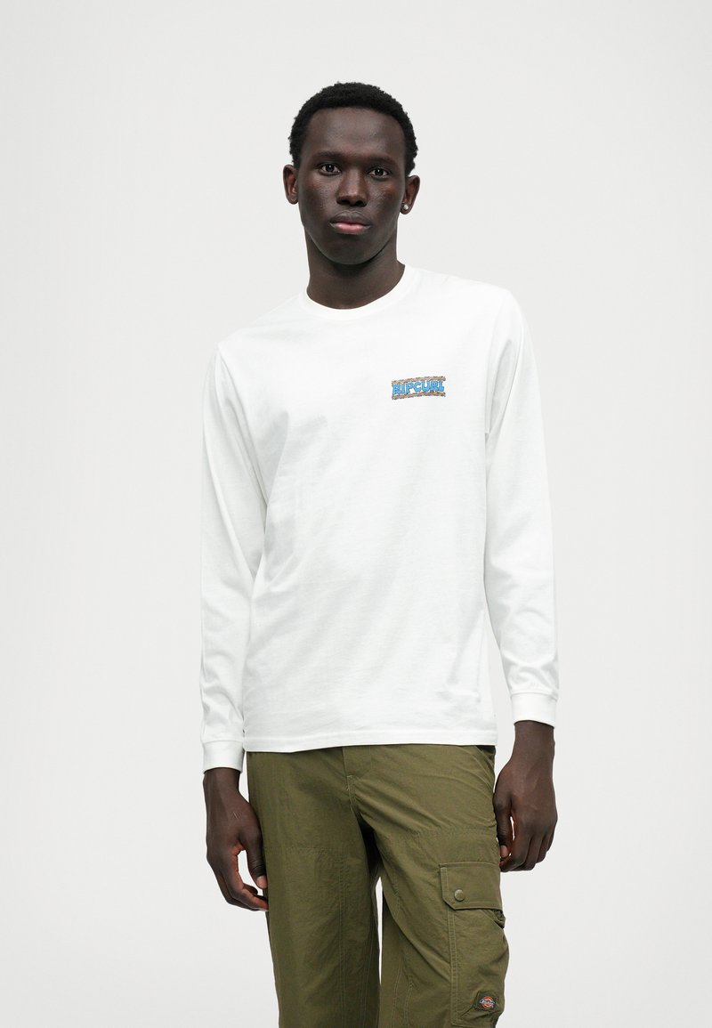 Langärmliges weißes Baumwoll-T-Shirt mit einem blauen und schwarzen Logo-Patch auf der Brust, kombiniert mit olivgrünen Cargo-Hosen.