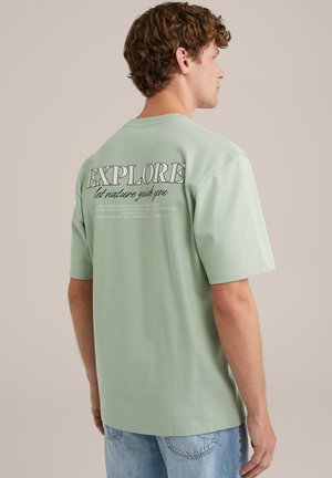 Camiseta de algodón verde con mangas cortas, con la frase "EXPLORA" y un gráfico de texto en la parte posterior. Diseño casual, corte relajado y textura suave.