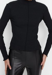 Schwarzes, geripptes Langarmoberteil mit hohem Kragen, das eine taillierte Passform und sichtbare Nahtdetails aufweist. Getragen mit schwarzen Leggings.