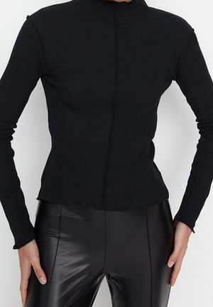 Haut noir à manches longues côtelé avec un col haut, présentant un design ajusté et des détails de coutures visibles. Porté avec des leggings noirs.
