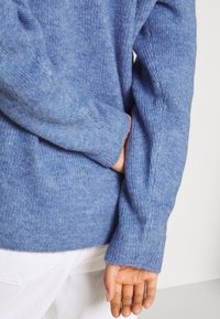Personne portant un pull en tricot bleu, une main rentrée à l'intérieur et l'autre le long du corps, portant un pantalon blanc.