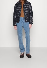 Black puffer jacket with a high collar, shiny texture, and front zip; paired with light blue jeans and chunky brown lace-up shoes.