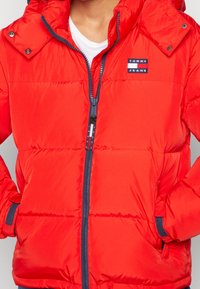 Red puffer jacket with a zip front, hood, and navy accents. Features quilted texture and a rectangular logo patch. Suitable for cold weather.