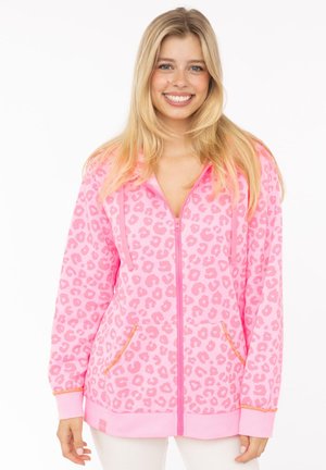Junge Frau, die lächelt, trägt einen rosa Kapuzenpullover mit Leopardenmuster und orange abgesetzten Taschen, steht vor einem weißen Hintergrund.