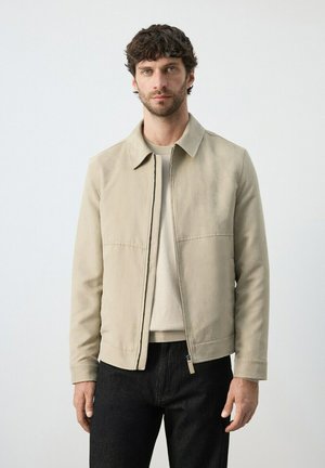 Man with short curly hair wearing a beige jacket over a light sweater and black pants, standing against a plain light background.