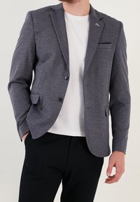 Mężczyzna w szarym żakiecie, pod którym ma białą koszulę z okrągłym dekoltem i czarne spodnie, z jedną ręką w kieszeni, a drugą swobodnie opartą przy boku.