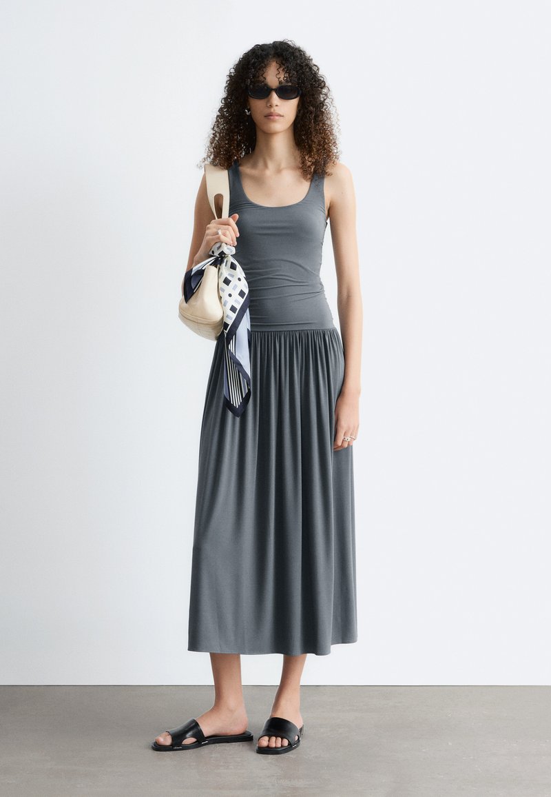 Femme aux cheveux bouclés portant des lunettes de soleil noires, une robe grise sans manches, des sandales slides noires, portant un sac à main crème avec un foulard noué.